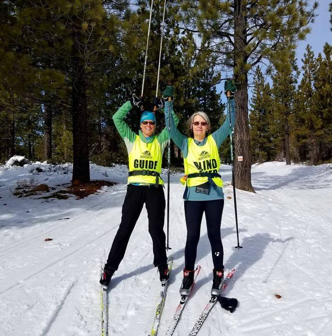 SFL promo photo - blind skier and guide standing on ski trail with arms raised in victory sign