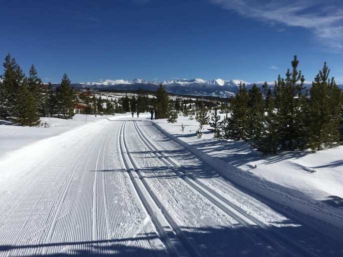 Two parallel set of cross country ski tracks heading down a gently sloping trail with mountains and trees in the background